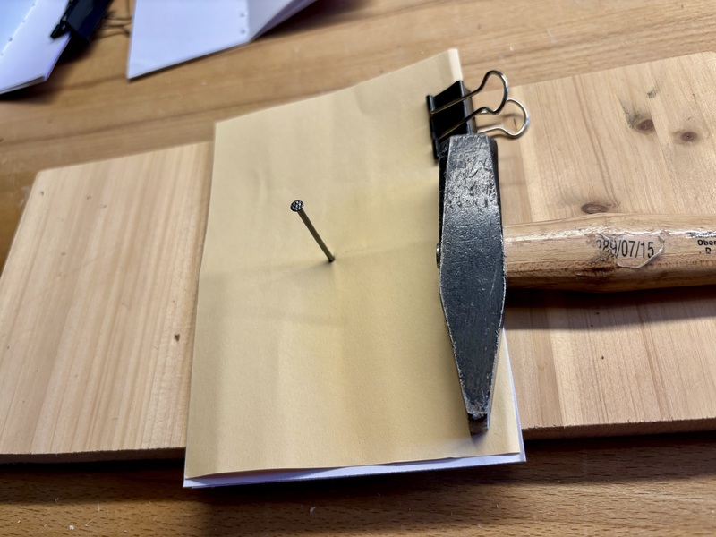 stacked sheets + cover sheet lying on a piece of wood. They're clipped together. A nail is sticking through the middle, and a small hammer is lying next to them.