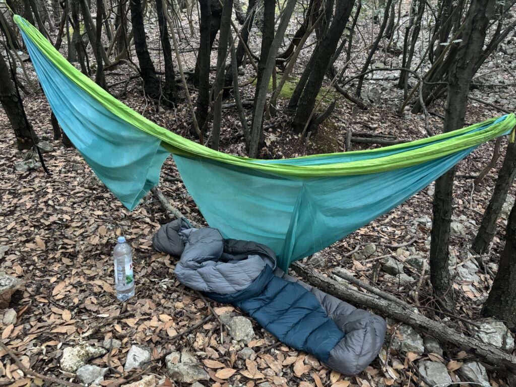 A sleeping bag lying on the ground, underneath a ripped hammock